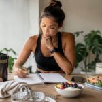 Mulher fisicamente ativa organizando alimentação e treino com caderno, tênis, shaker, fita métrica e refeição saudável sobre a mesa.